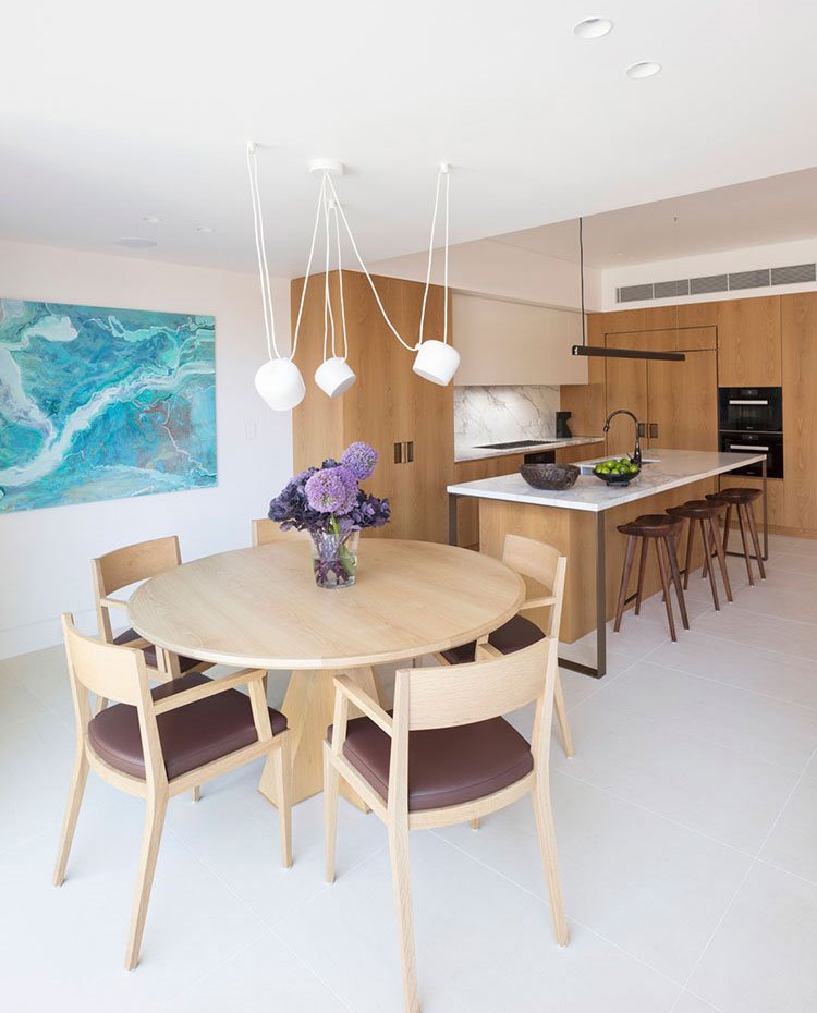 Mesa de comedor y sillas de madera, luminaria suspendida con varias unidades, cocina abierta al fondo