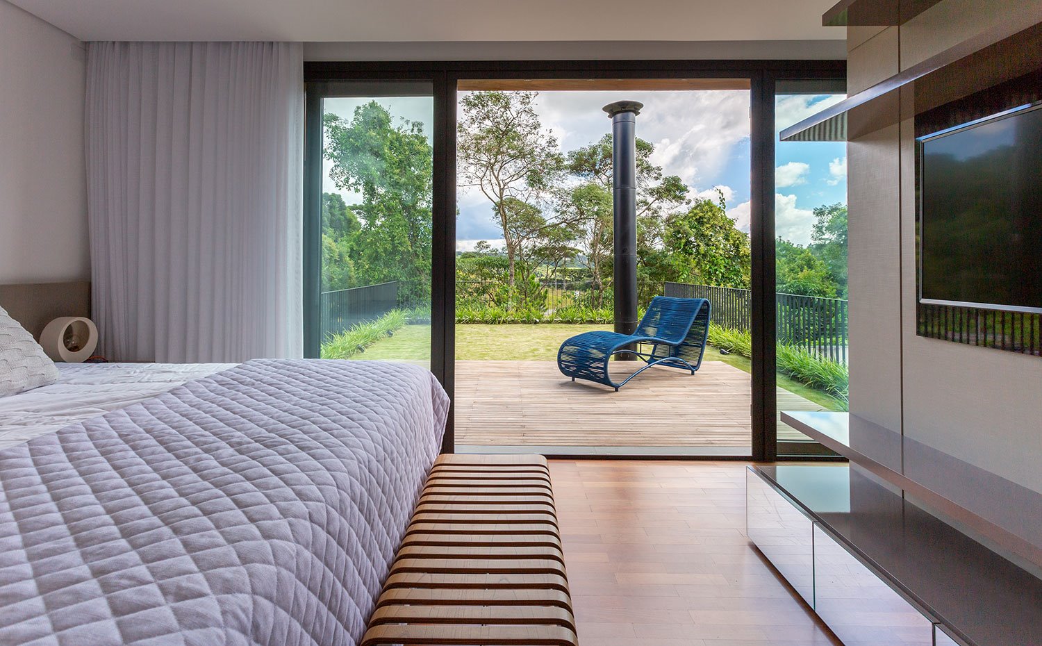 Dormitorio principal con bancada de madera, mueble frente a cama para televisión, terraza con plataforma de madera y chaise-longue azul