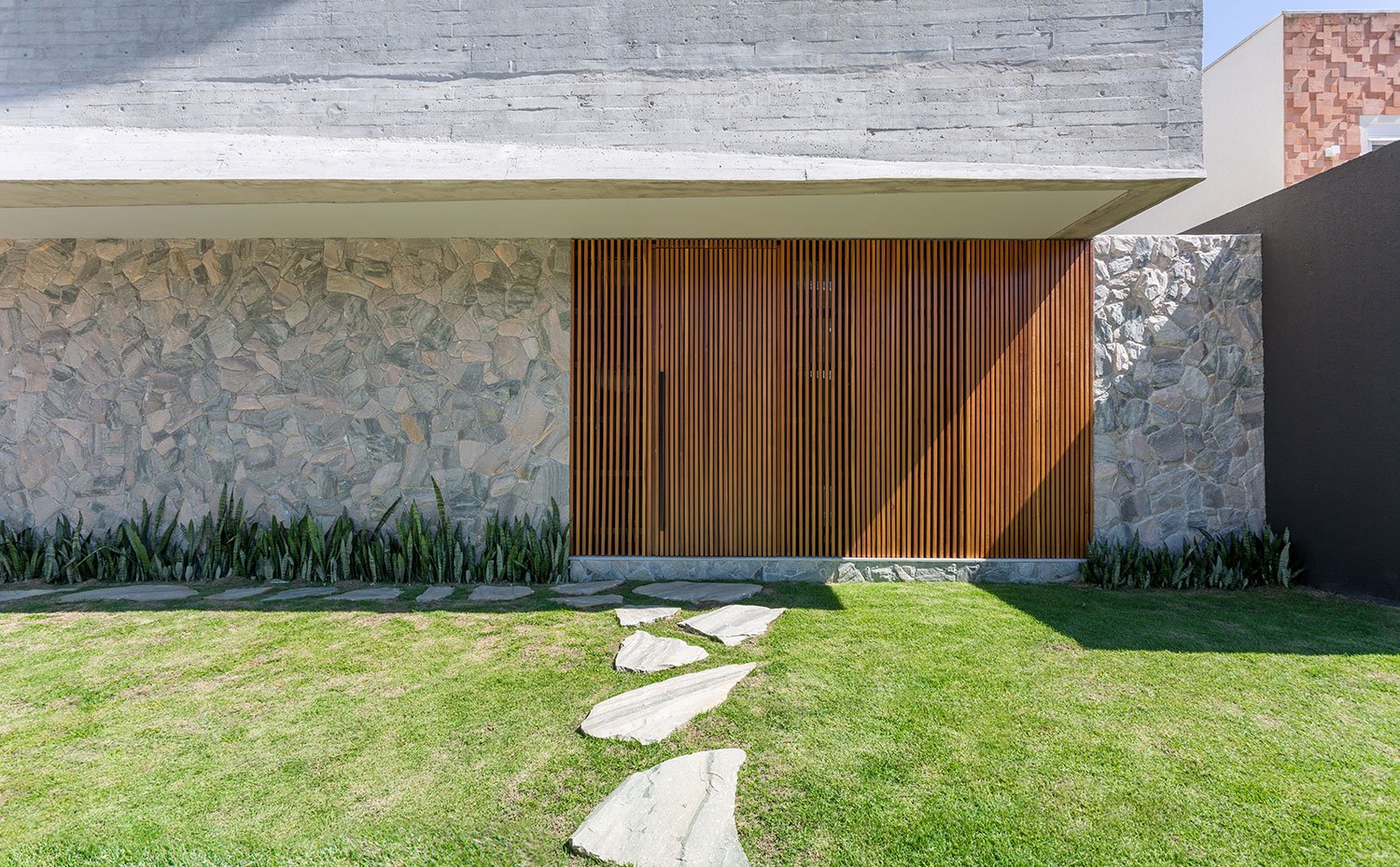 Entrada vivienda oculta tras frente alistonado de madera en hueco de fachada de piedra y hormigón