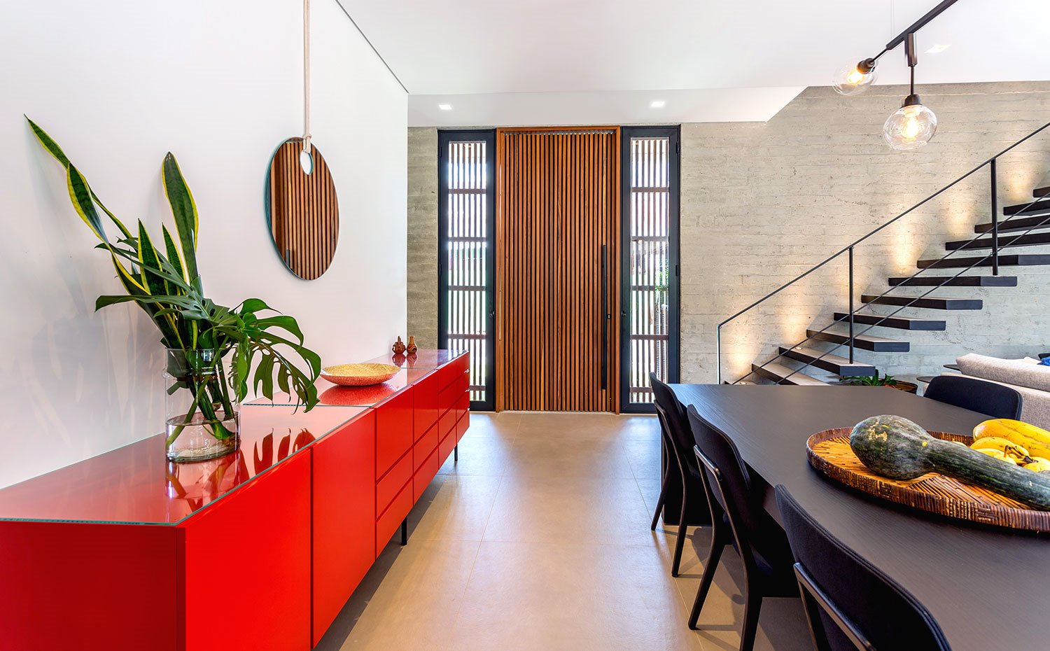 Mueble contenedor lacado en rojo, mesa de comedor con sillas en negro, puerta de acceso en cristal y madera, frente de pared de hormigón con escaleras en negro