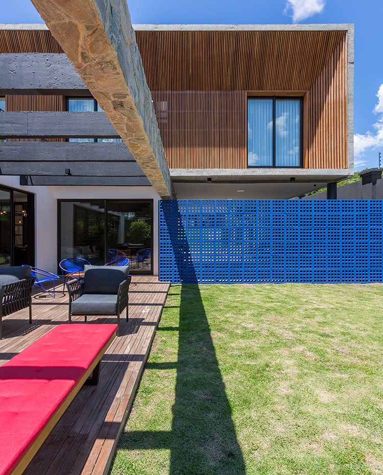 Pavimento con listones de madera sobre zona ajardinada, celosía en azul, butaca en negro, fachada en piedra y madera