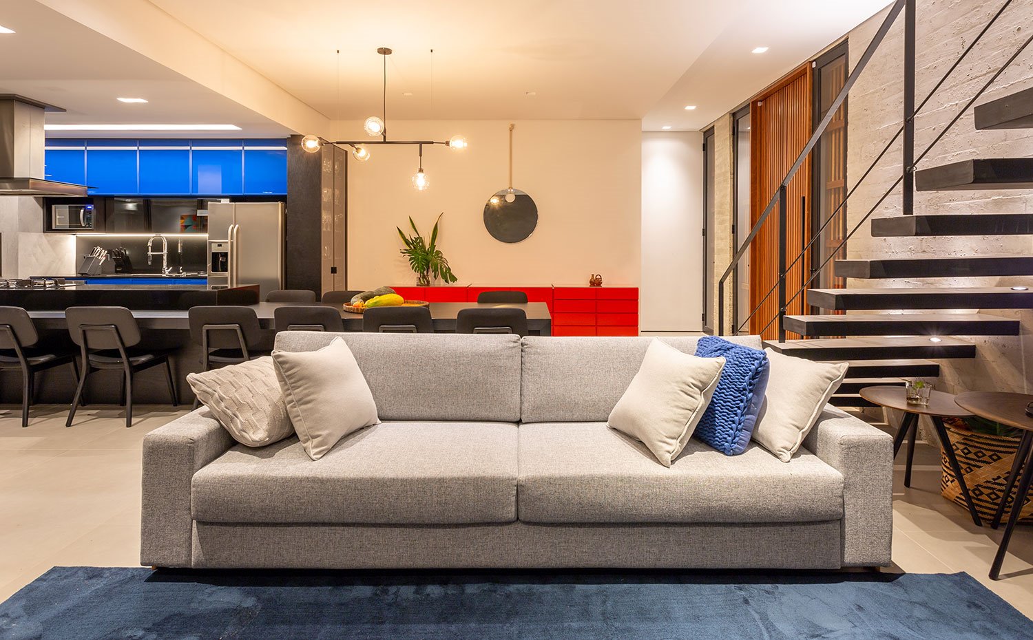 Salón con sofá gris y alfombra azul sobre pavimento en gres porcelánico, mesas auxiliares con sobre de madera bajo escaleras