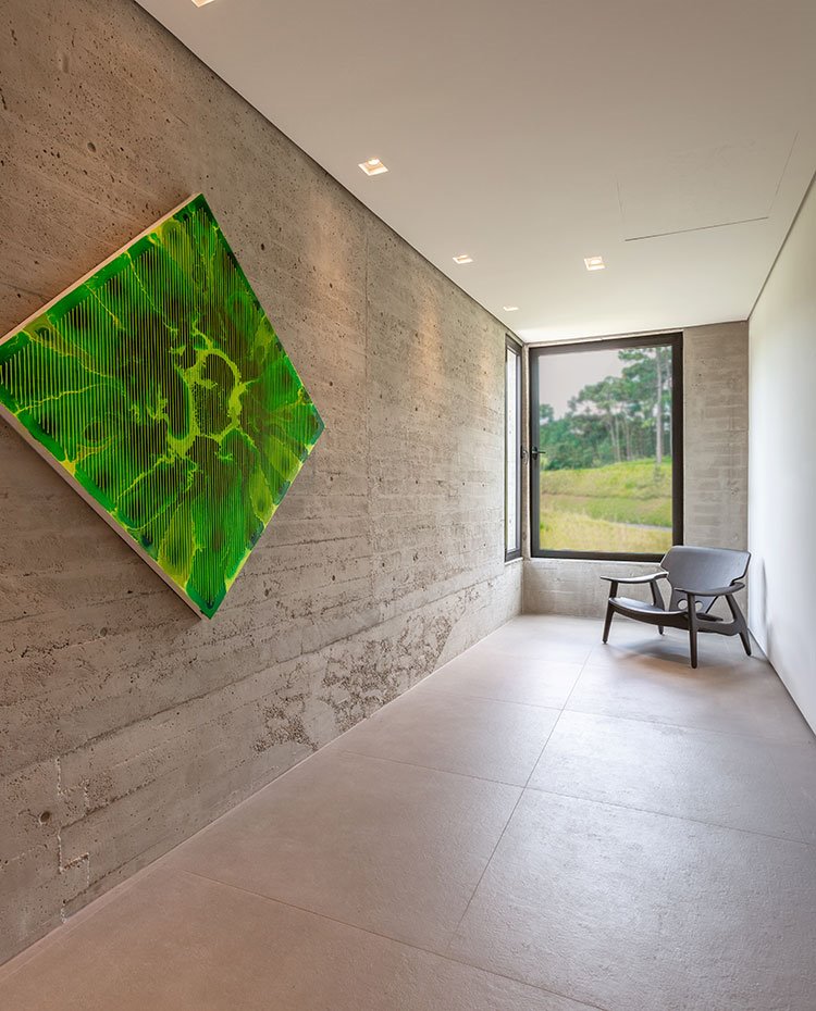 Zona de paso nivel superior, con obra artística en verde sobre pared de hormigón, butaca de madera y aperturas en fachada en cristal transparente