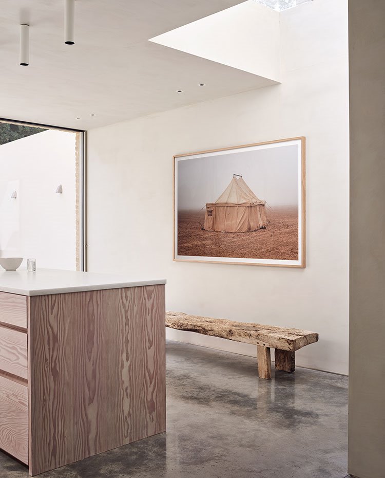 Isla de trabajo en cocina en madera veteada y encimera blanca, suelo de hormigón y bancada en madera natural, focos blancos en techo y lucernario