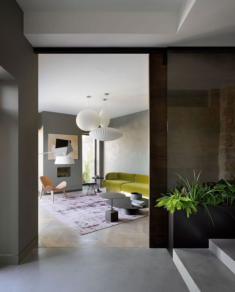 Acceso a salón con sofá en verde pistacho, butaca de madera, mesas en negro y alfombra en lila.