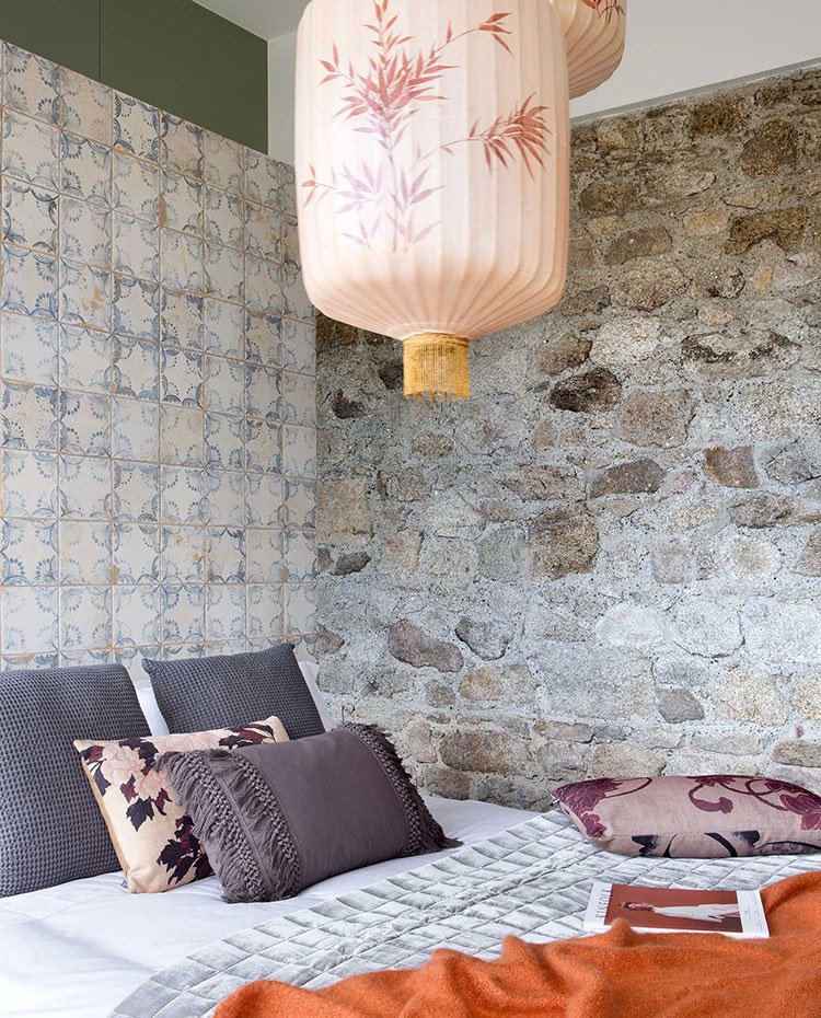 Dormitorio con cojines en gris, cabecero con baldosas antiguas recuperadas, pared de piedra y luminaria suspendida de inspiración oriental