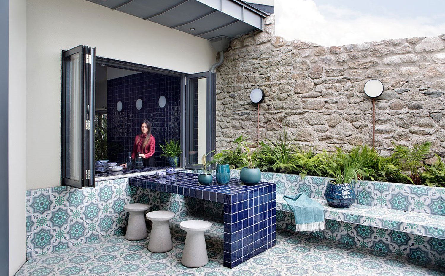 Patio exterior con ventana y mesa en comunicación con el interior, mesa de obra azil, taburetes en gris y bancada recubierta de baldosas