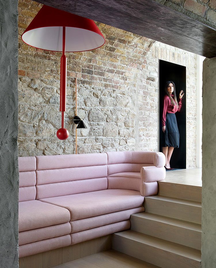 Sofa de formas curvilíneas en rosa junto a escalera, luminaria suspendida en rojo, pared de ladrillo y piedra vista