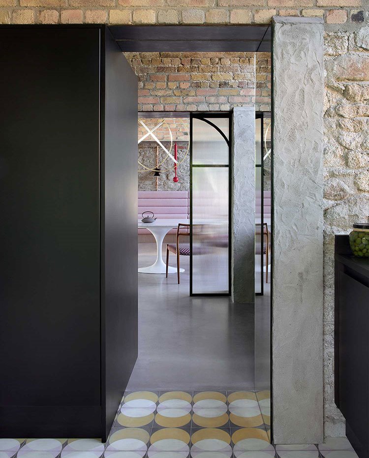 Suelo con motivos geométrios y puerta de acceos en negro en contraste con pared de piedra, suelo de hormigón contínuo hacia comedor