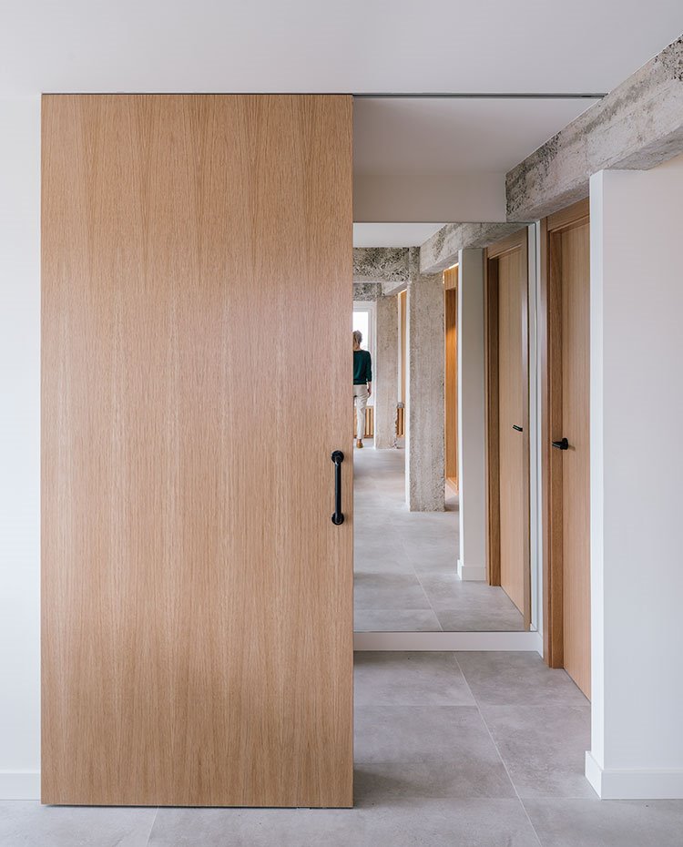 Puerta corredera con tirador negro, suelo porcelánico gris y paredes blancas con viga de obra