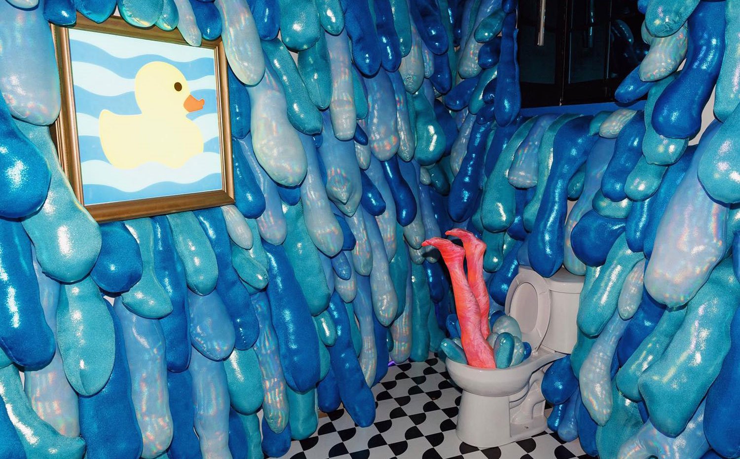 Inodoro con camas de maniquí, paredes a modo de cojines en azul y cuadro con pato.
