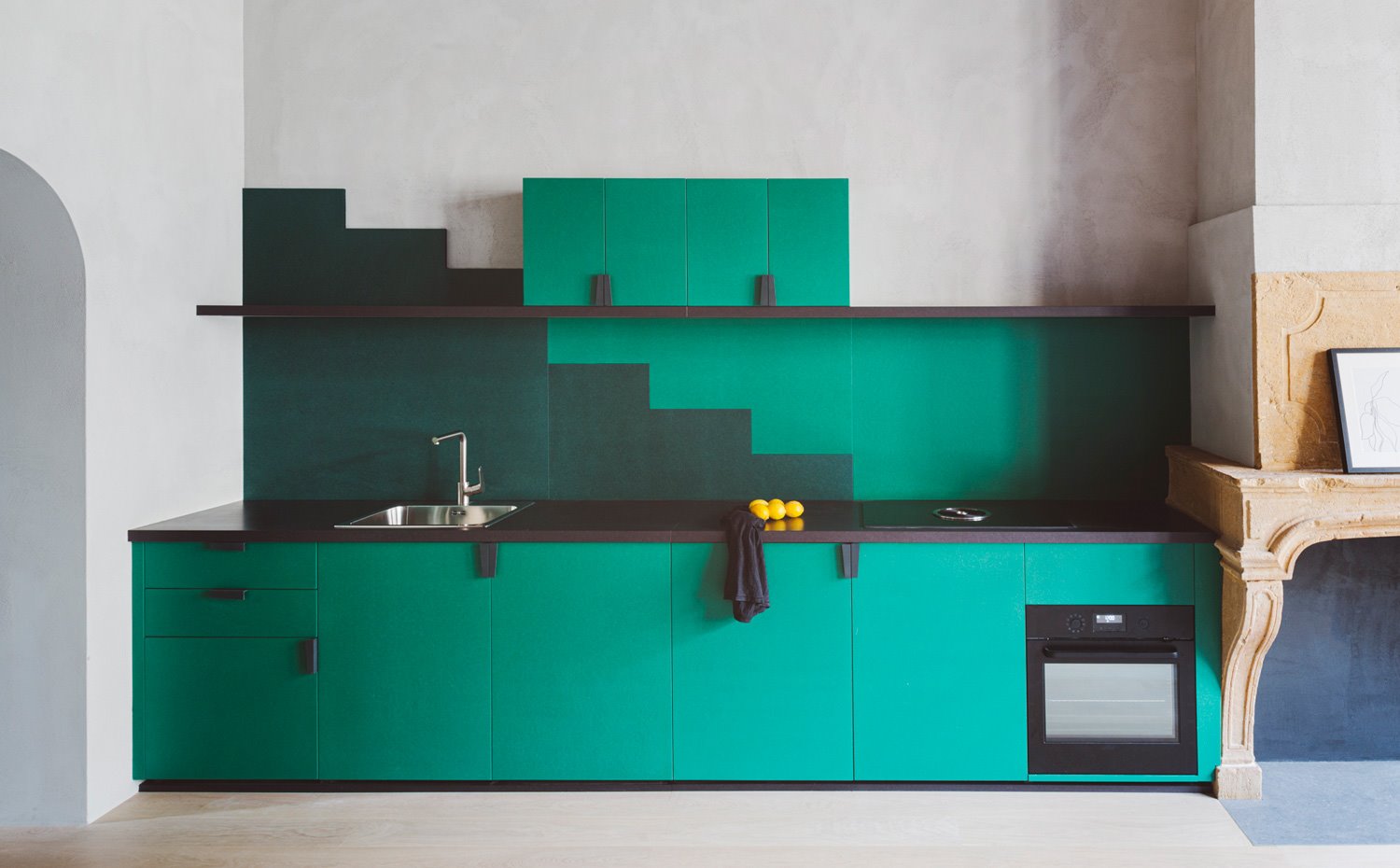 Cocina con frentes de armarios en verde, revestimiento bitono fregadero de acero y electrodoméstico en negro, chimenea antigua en mármol beige