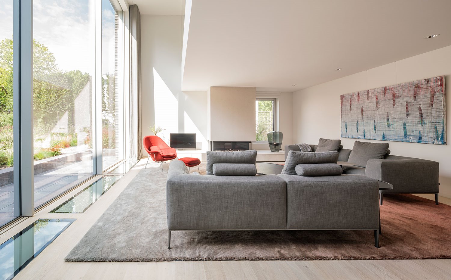 Salón con sofás en tonos grises, alfombra en tonos piedra y butaca y pouf en rojo