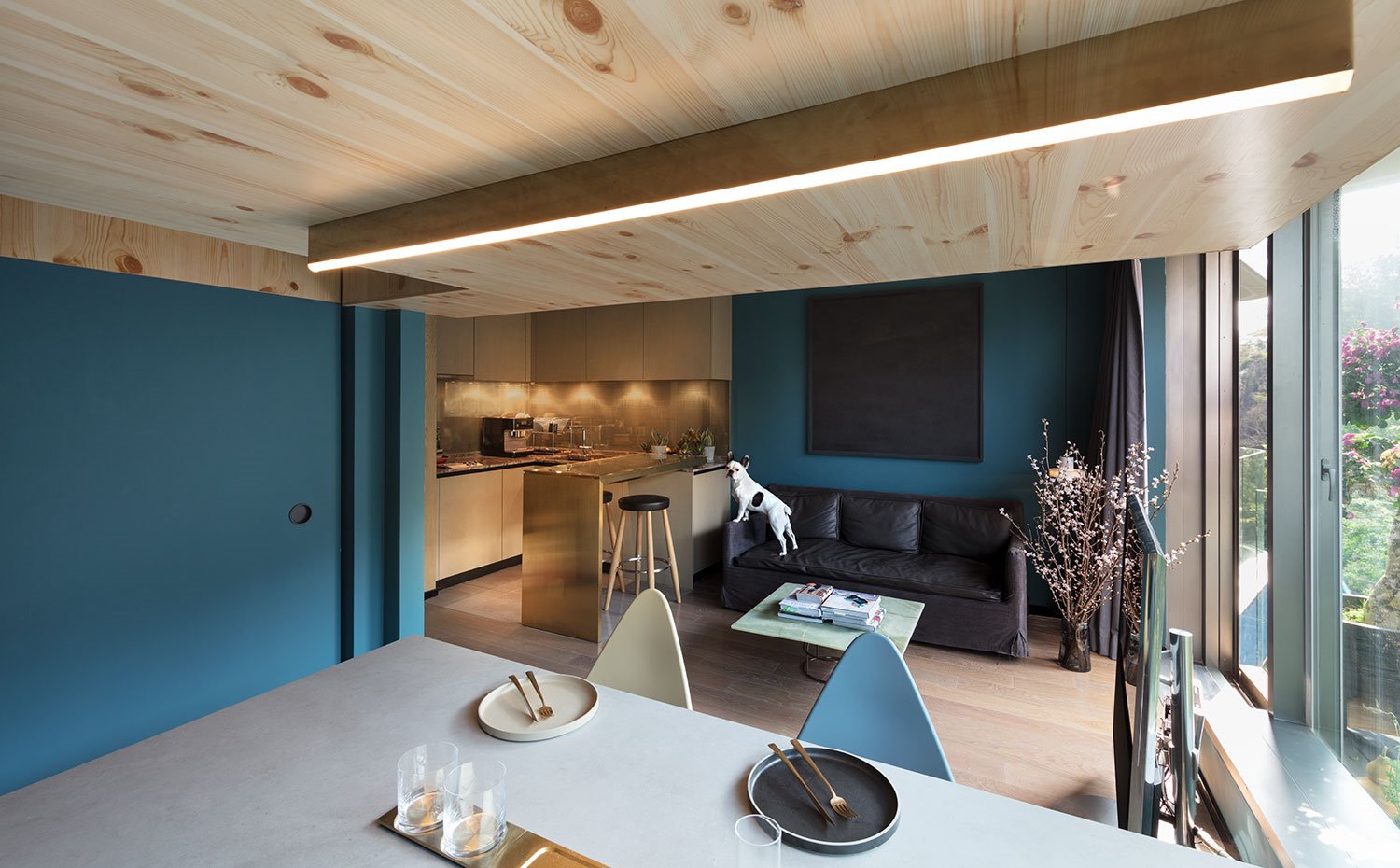 Cocina y comedor abiertos junto a comedor, paredes pintadas de azul con revestmiento de madera, sofá en negro