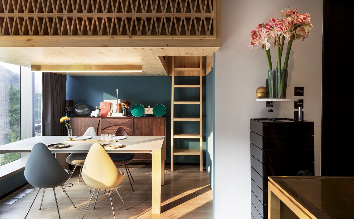 Mesa comedor de madera y mueble contendor de madera oscura, sillas en gris y crema, estante con jarrón con flores