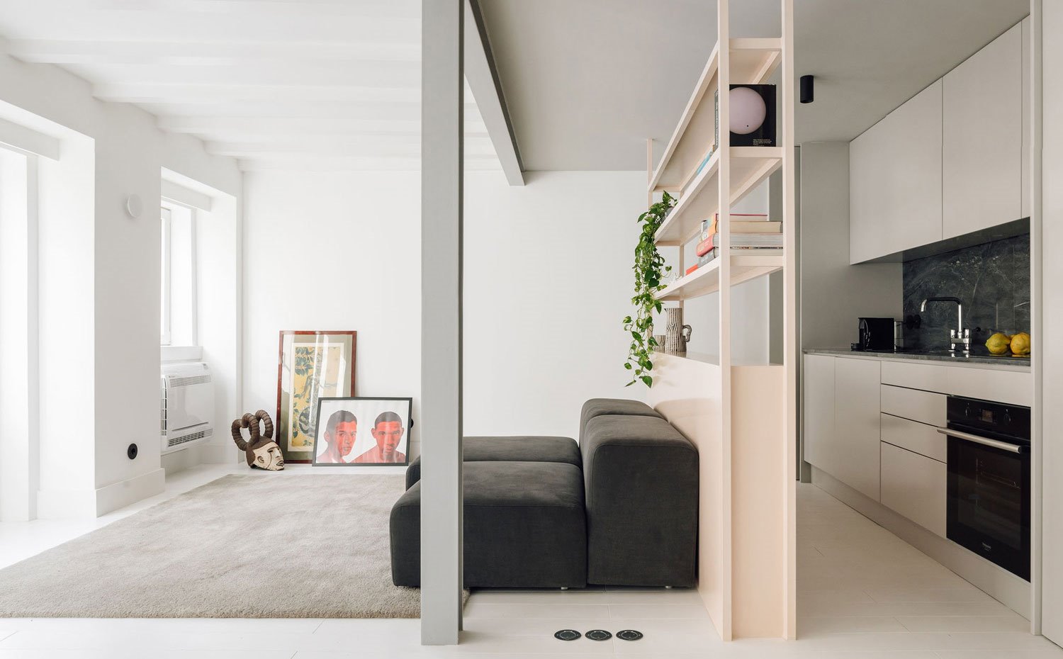 Salón y cocina separados por módulo a modo de estante abiertos, sofá bajo gris oscuro, alfombra y vigas en gris. 