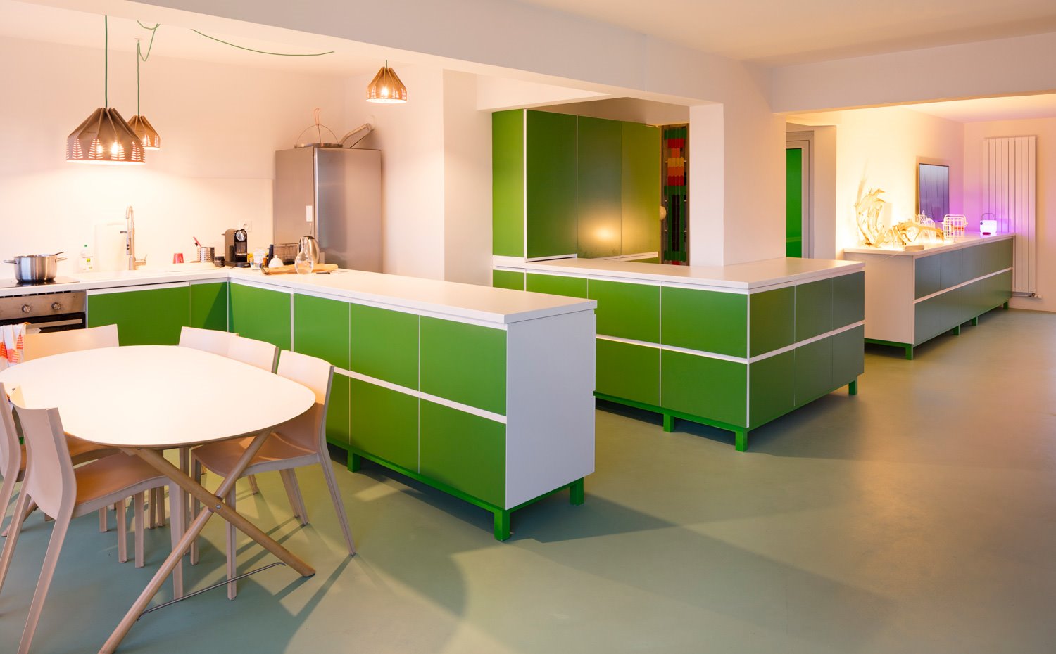 Comedor con sillas en beige y cocina con muebles bajos en verde dispuesto en forma de L, armarios en pared verdes