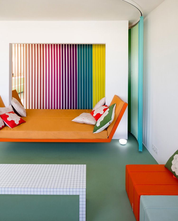 Sofa-cama en naranja, mesa blanca, poufs en naranja y gris y suelo contínuo en verde