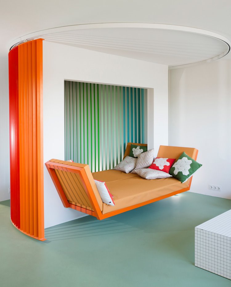 Sofá narranja con cojines de colores, estructura curva de acero en diferentes colores, suelo contínuo en verde