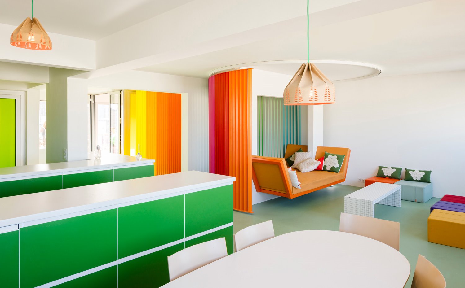 Zona abierta, con módulos de almacenaje en verde, luminarias suspendidas con pantallas troqueladas, poufs de colores entorno a mesa de centro rectangular blanca