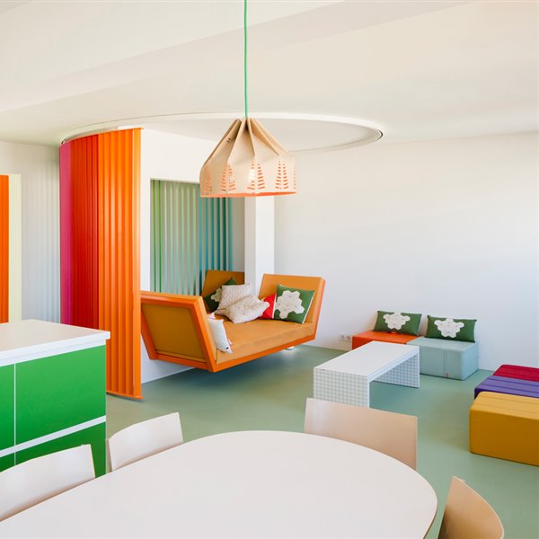Zona abierta, con módulos de almacenaje en verde, luminarias suspendidas con pantallas troqueladas, poufs de colores entorno a mesa de centro rectangular blanca