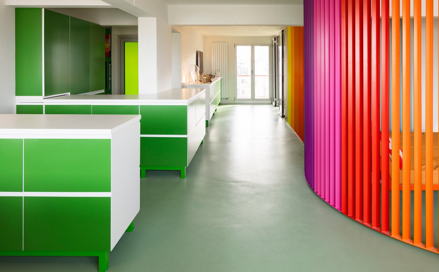 Zona de paso entre cocina, con mobiliario en verde, y biombo en naranja, rojo, rosa y lila