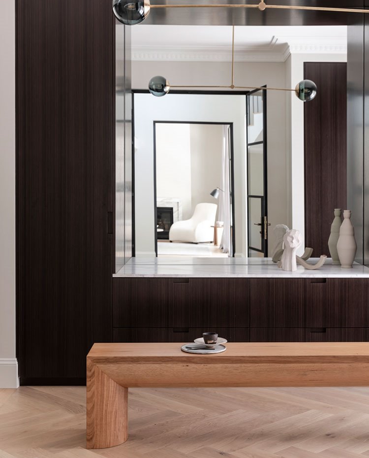Bancada baja de madera con complemento de cerámica, mueble de almacenaje en madera oscura, con sobre en blanco con la parte central revestida de espejo, molduras en techos y suelo de madera