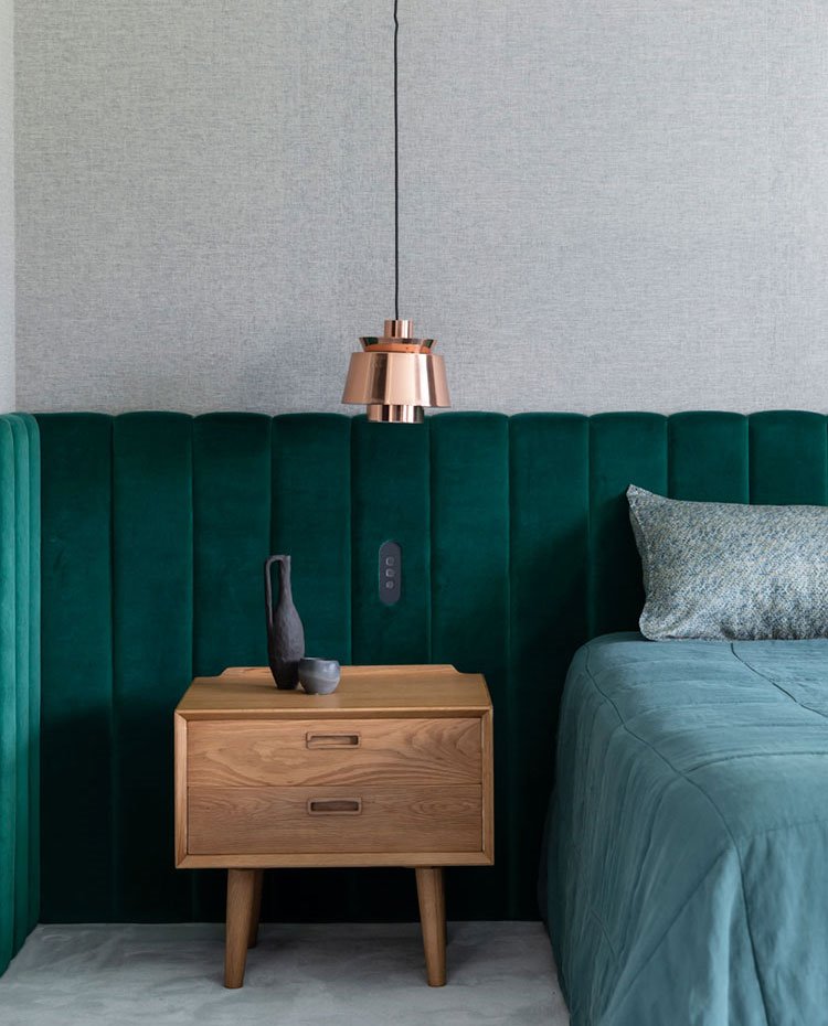 Cabecero a lo largo de todo el perímetro de la pared del dormitorio acolchado en verde botella, mesilla de madera y luminaria suspendida con pantalla en acabado latón