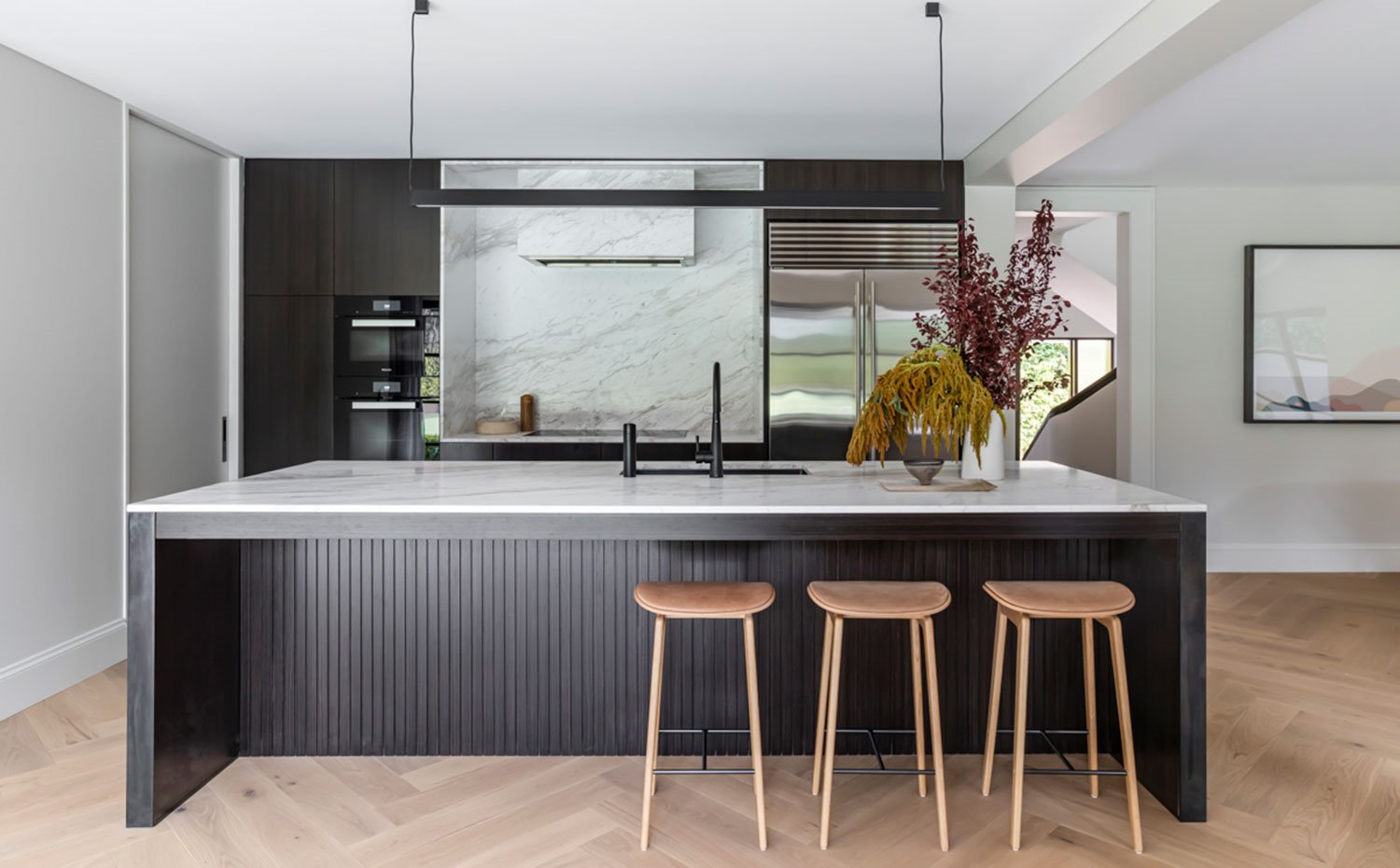 Cocina abierta con mobiliario en madera oscura, frente al suelo de madera clara en zig-zag. Hueco de isla de trabajo para taburetes, encimera en mármol blanco