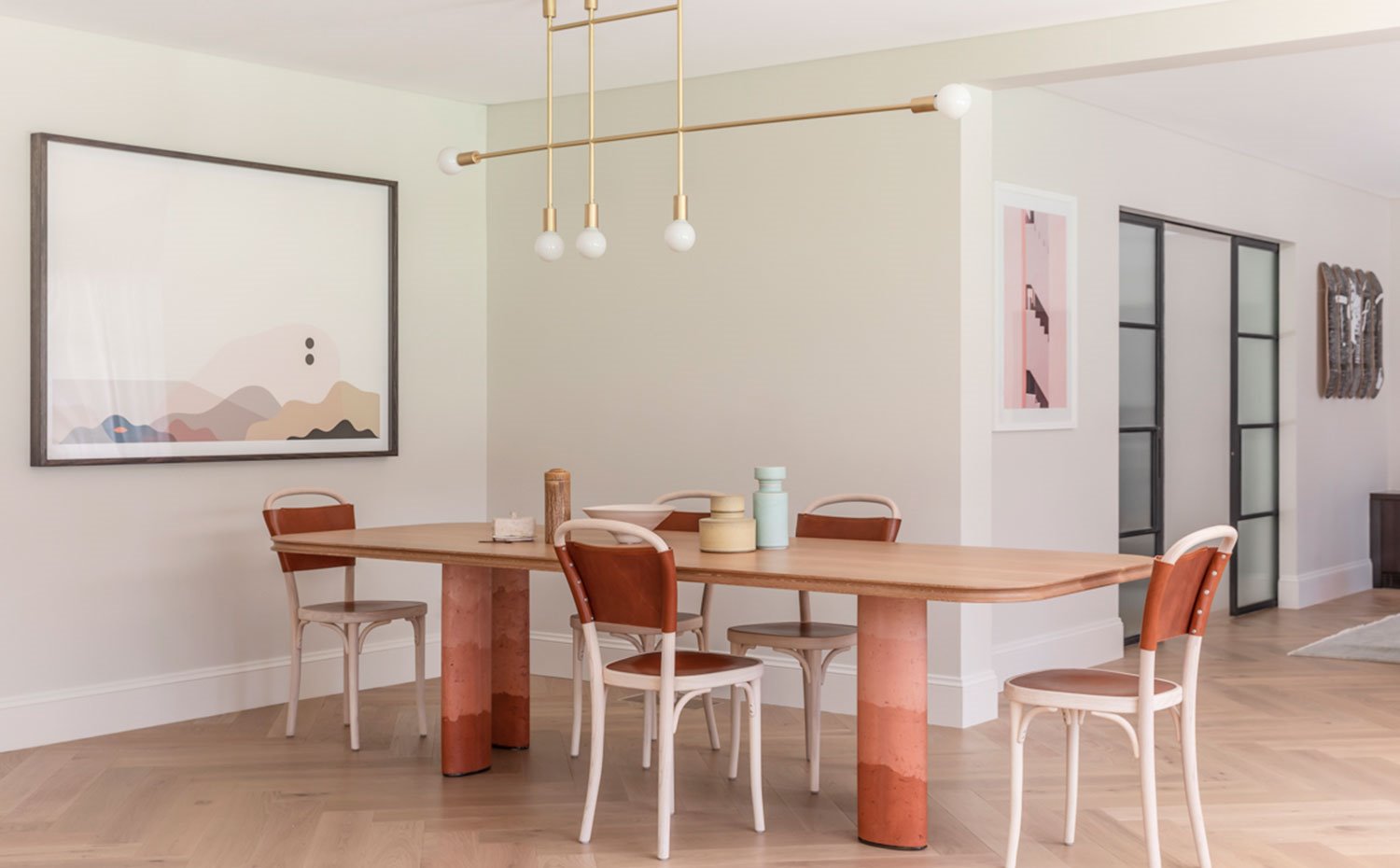 Comedor con sillas en madera y respaldo de piel, y amplia mesa con sobre de madera y patas con detalle degradado. Luminaria suspendida dorada y suelo de madera