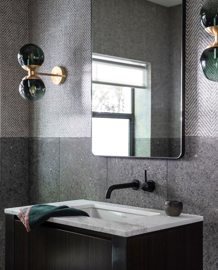 Cuarto de baño con espejo de cantos curvos, apliques de cristal verde y dorado, revestimiento en gris con motivos en zig-zag, encimera de mármol blanco, grifería en negro y módulo de madera.