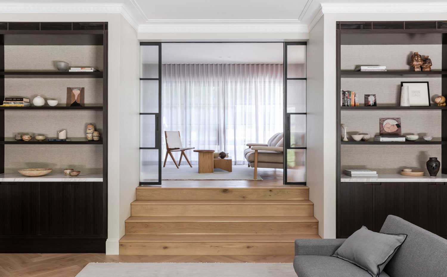 Estanterías en madera oscura flanqueando la zona de paso con escaleras que conducen a otra sala de estar elevada, sofá en gris, puertas correderas de cristal y perfilería oscura