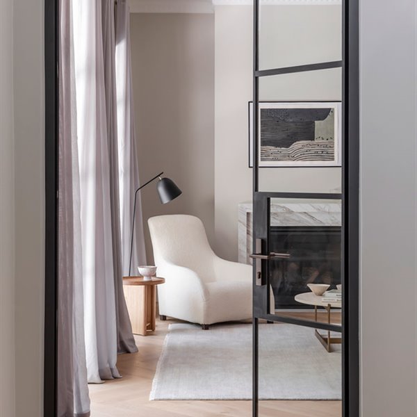 Puerta de cristal hacia salón con perfilería en negro, suelo de madera, alfombra blanca y butaca junto a cumenea con marco de mármol