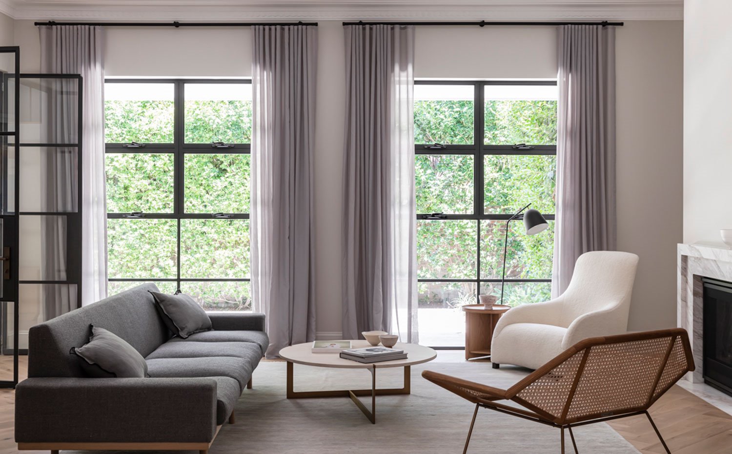 Salón con amplios ventanales provistos de cortinas grises, butaca cruda, sofá ris y asiento con estructura perforada, mesa de centro circular de madera
