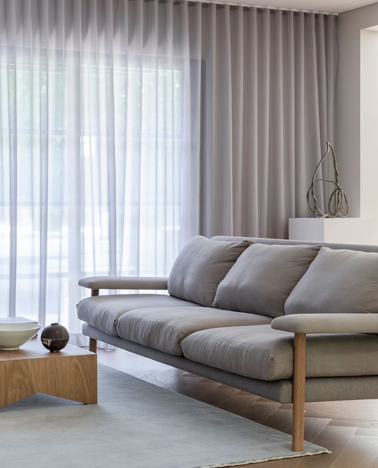 Salón con sofá gris claro con estructura de madear, mesa de centro cuadrada en madera sobre alfombra en crudo, cortinas grises corridas y sueno de madera
