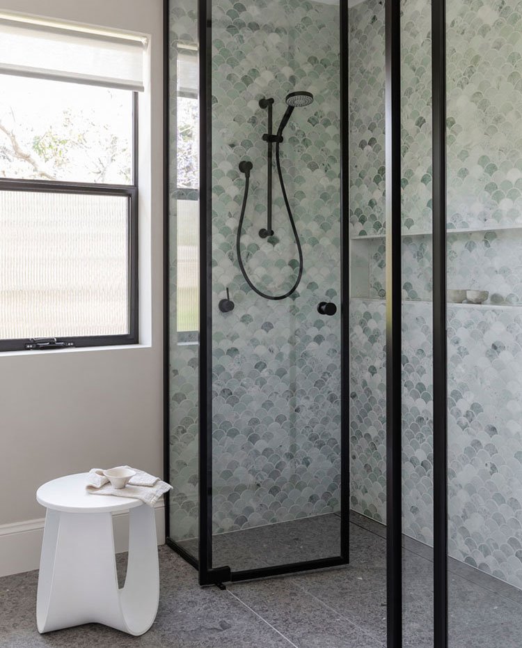 Zona de ducha con cerramiento acristalado a modo de hojas abatibles, perfilería en negro, revestimiento de ducha en verde con hornacina de obra, taburete en blanco