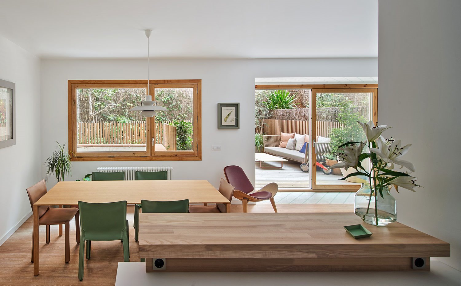 Voladizo de cocina en madera sobre encimera, mesa de comedor en madera con sillas en verde y marrón, butaca de madera con revestimiento en granate