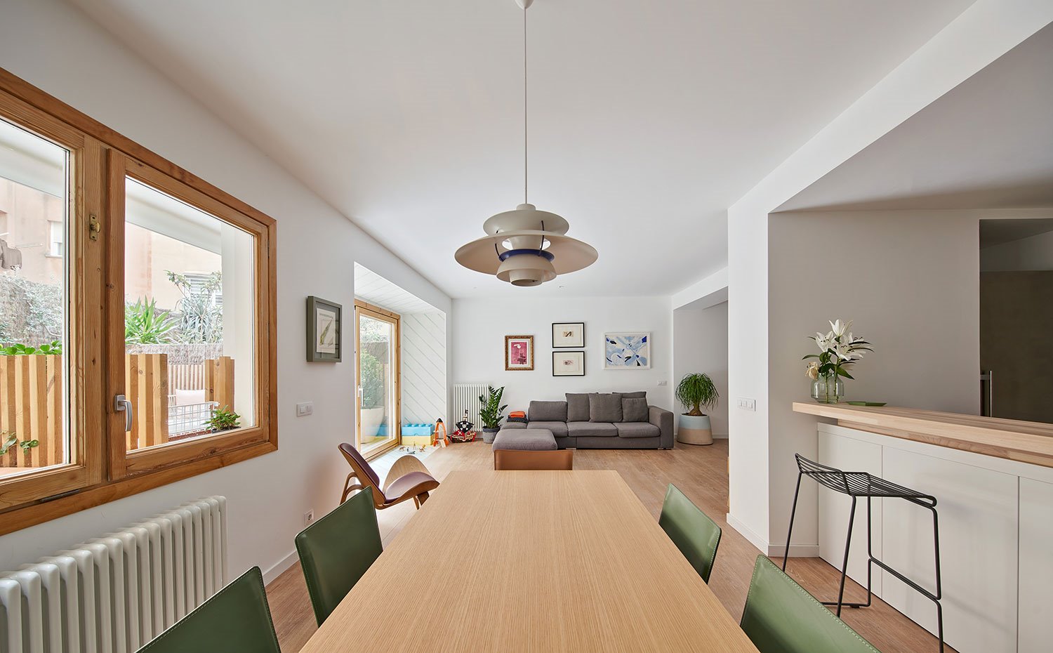 Zona comedor con mesa de madera y sillas en verde, luminaria suspendida vintage, taburete negro junto a zona office de cocina semiabierta