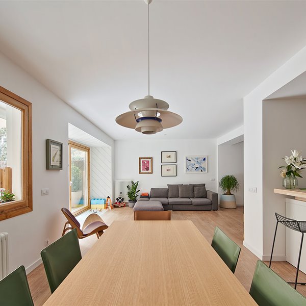 Zona comedor con mesa de madera y sillas en verde, luminaria suspendida vintage, taburete negro junto a zona office de cocina semiabierta