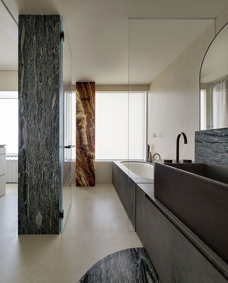 Cuarto de baño con columnas y revestimientos en marmol, encimera de piedra gris, cerramiento de cristal