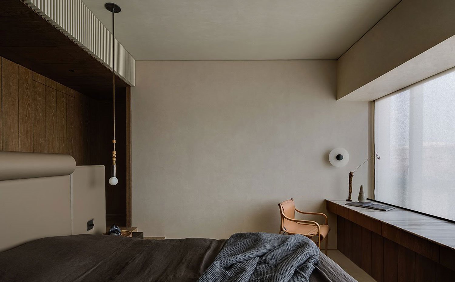 Dormitorio con cabecero revestido de piel, ropa de cama en gris, luminaria suspendida de techo, zona de trabajo en pies de cama