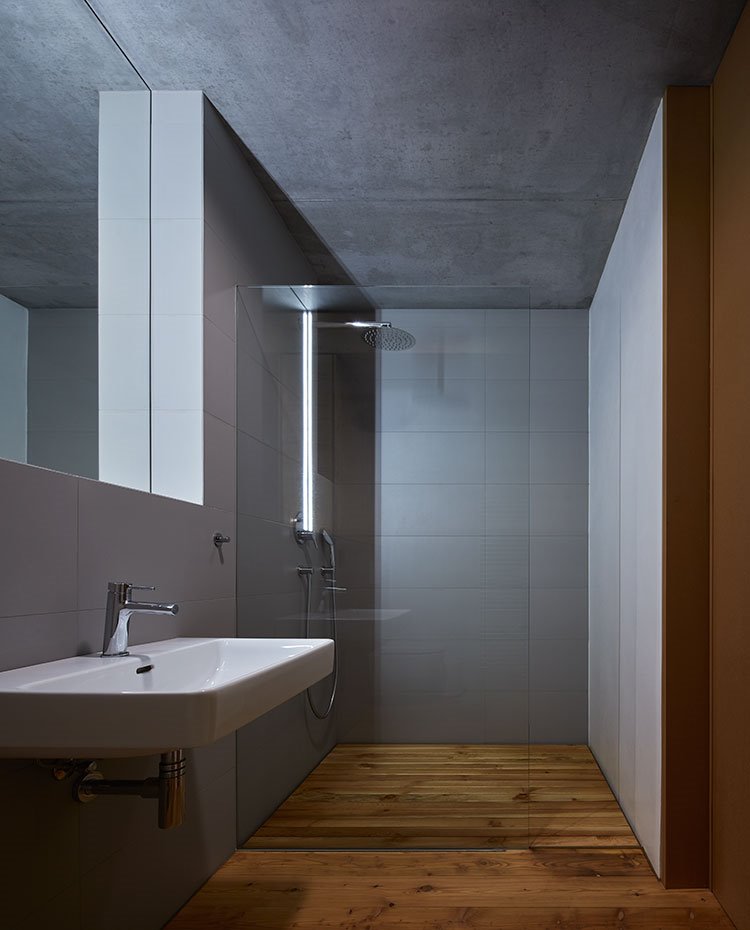 Cuarto de baño con suelo de madera, revestimiento en gris y techo de hormigón, hoja de cristal transparente en ducha