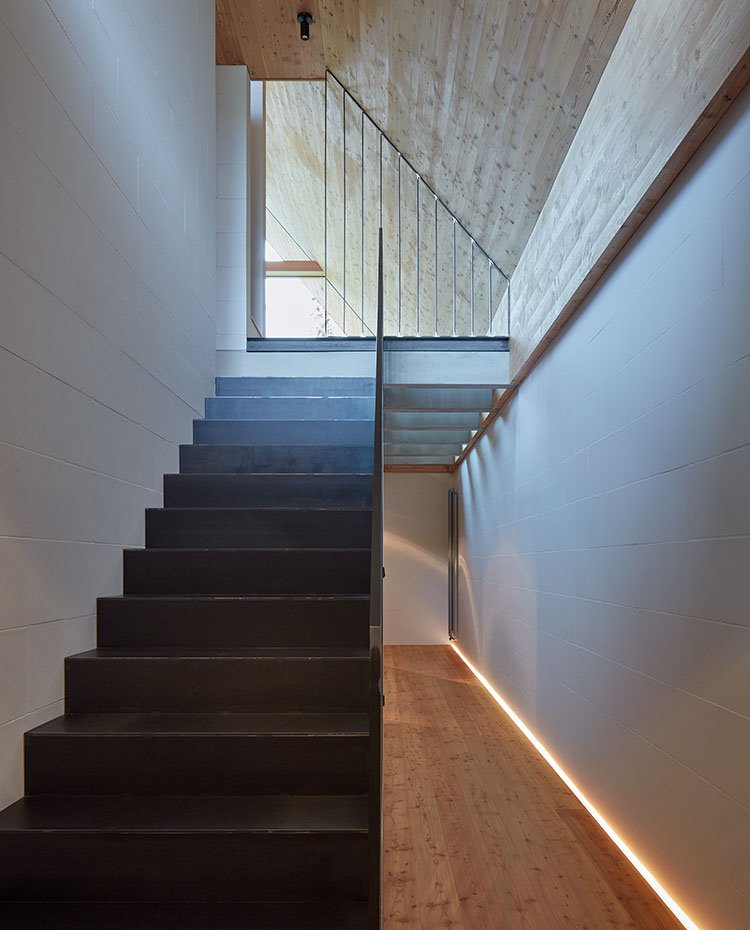 Escaleras entre los dos niveles de la casa, paredes blancas, parte superior y cubierta revestidas de madera, luminaria a modo de riel en el suelo 