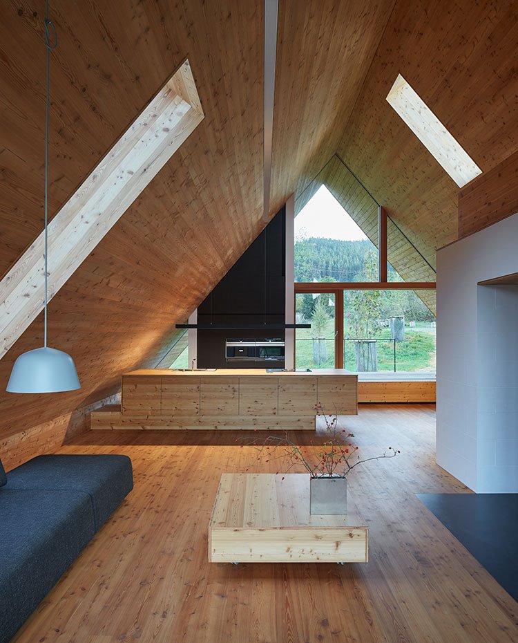 Espacio diáfano recubierto de madera con zona de cocina abierta y salón, mesa de centro en madera, sofá en gris y luminaria suspenda en gris claro