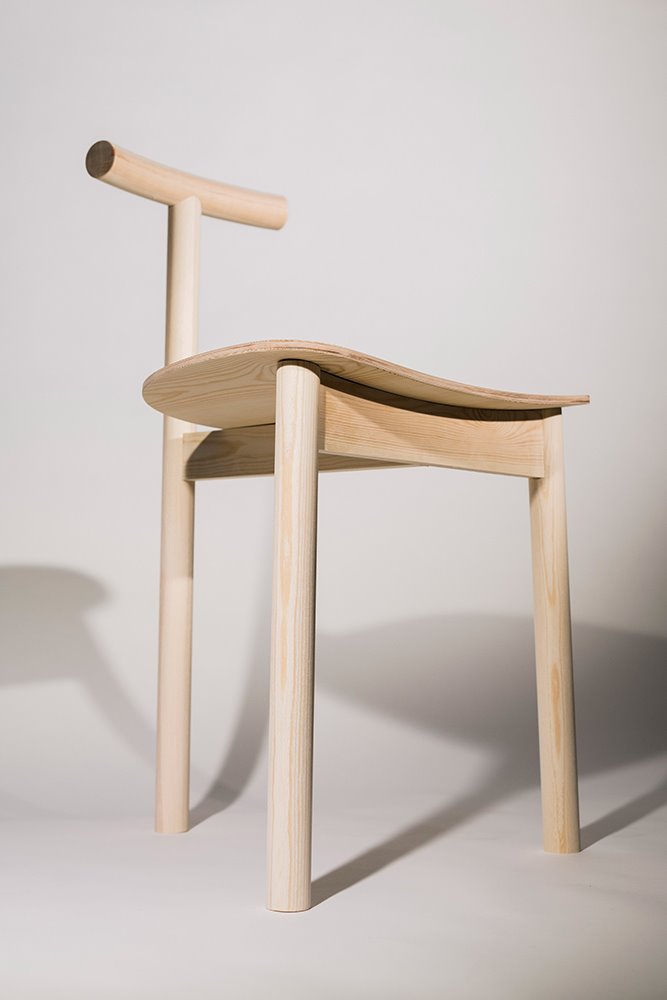 La silla destaca por su asiento ligeramente curvado apoyado sobre tres patas, una de las cuales se prolonga en un respaldo mínimo. 
