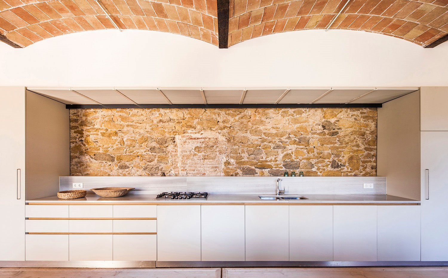 Cocina abierta, con salpicadero a media altura y pared de piedra, sin muebles altos, frentes en blanco con tiradores integrados