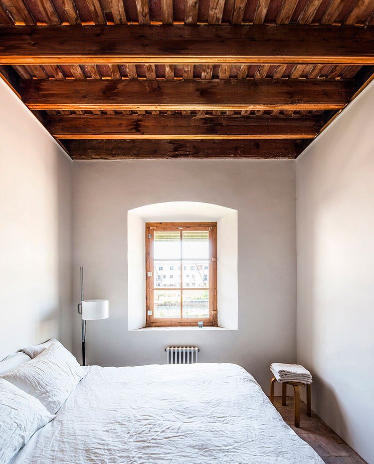 Dormitorio en blanco, con ventana de marcos de madera, luminaria de pie, techos de madera