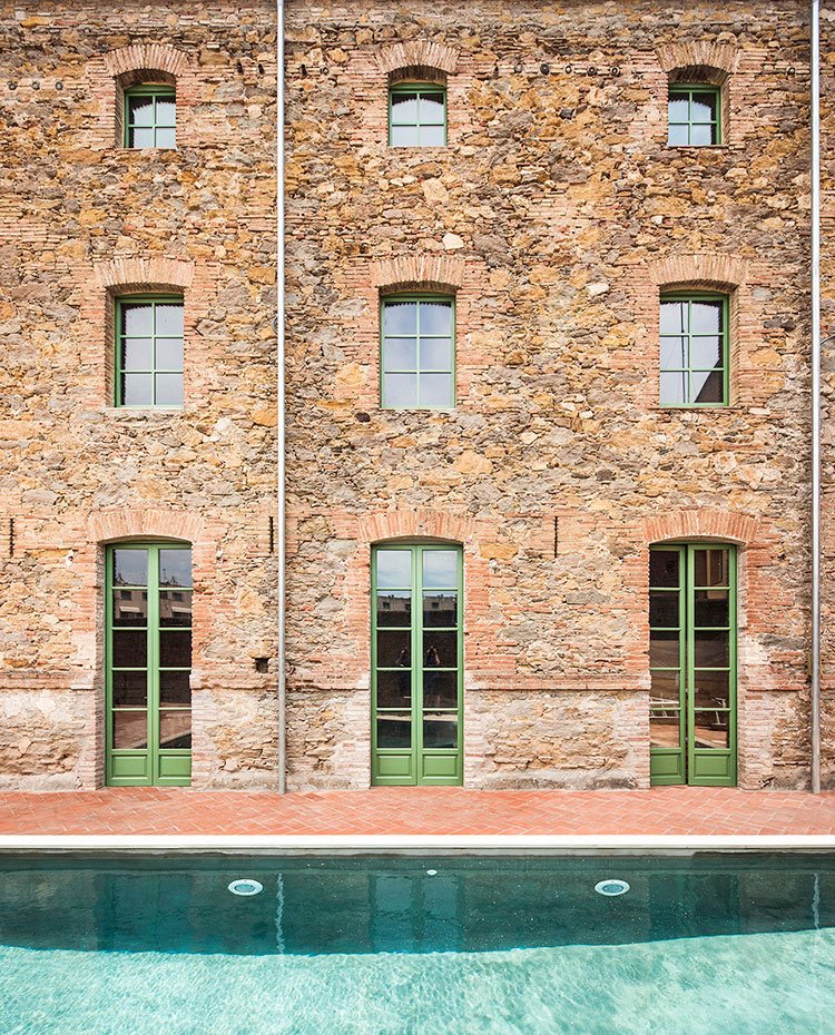 Fachada exterior de vivienda de piedra con aperturas con arcos de ladrillo, cerramientos con perfiles en verde, piscina en primer término