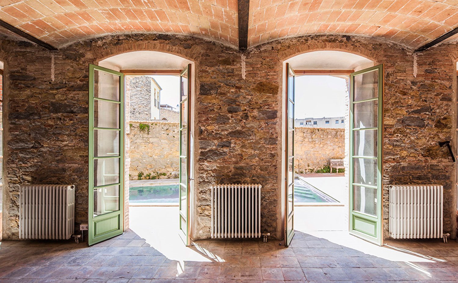 Interior vivienda con volta catalana, pared de piedra, aperturas a exterior con piscina en terraza, radiadores blancos en pared