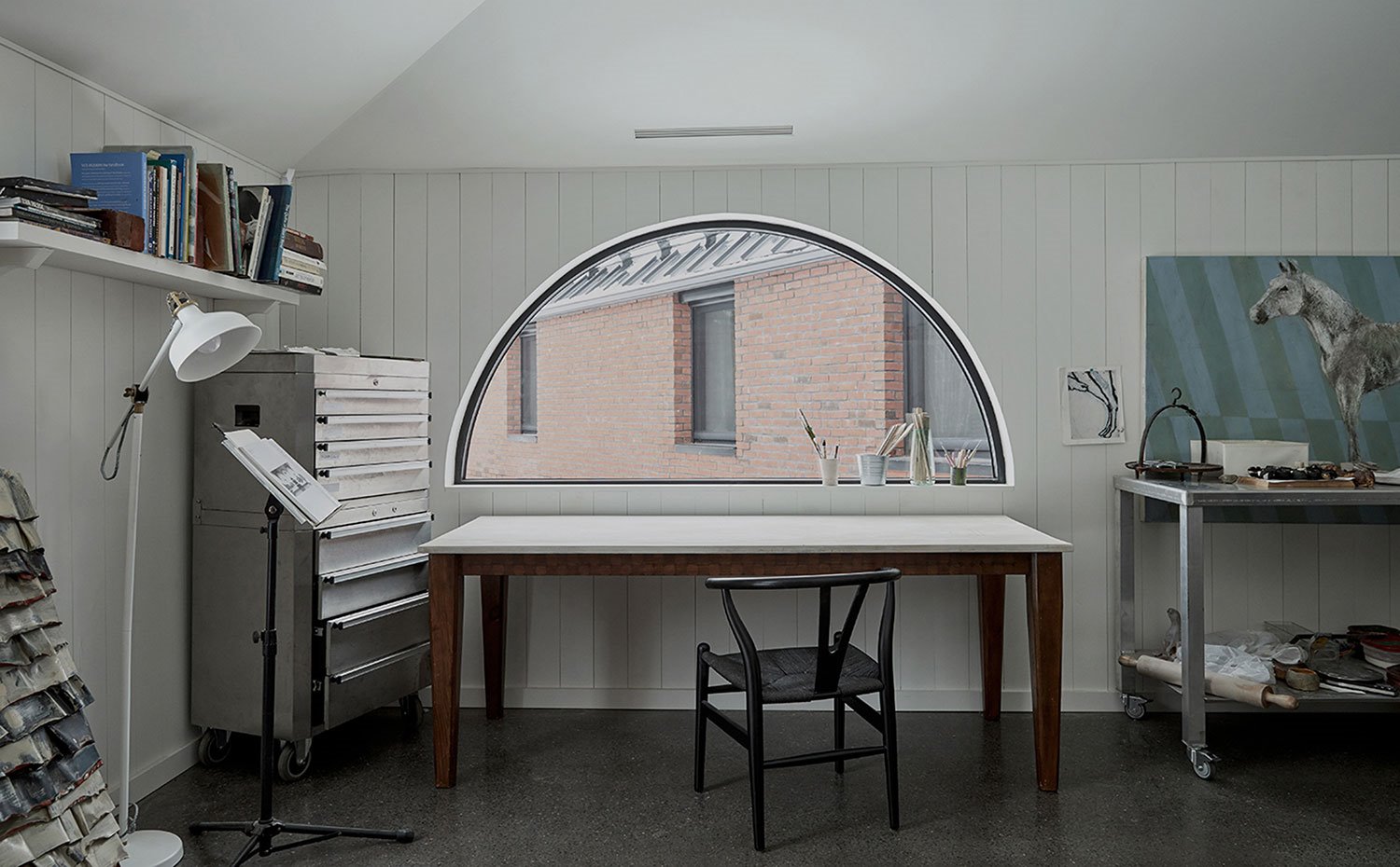 Estudio de trabajo con pared revestida de madera pintada de blanco, mesa y silla vintage, ventana en forma de semicírculo